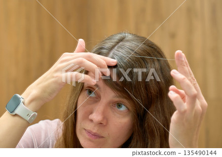 Woman examining hair roots and gray strands while looking at mirror Woman examining hair roots and gray strands while looking at mirror 135490144