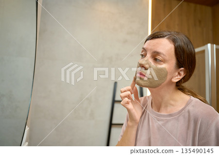 Woman standing in bathroom and applying clay face mask while looking at mirror Woman standing in bathroom and applying clay face mask while looking at mirror 135490145