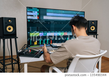 Skilled sound engineer working in home recording studio, using digital audio workstation software on large monitor and midi keyboard to compose and mix music. Skilled sound engineer working in home recording studio, using digital audio workstation software on large monitor and midi keyboard to compose and mix music. 135490344