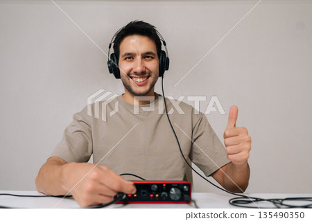 Cheerful audio engineer wearing headphones showing thumbs up gesture while adjusting audio interface, focusing on music production, sound recording, mixing audio and mastering tracks. Cheerful audio engineer wearing headphones showing thumbs up gesture while adjusting audio interface, focusing on music production, sound recording, mixing audio and mastering tracks. 135490350