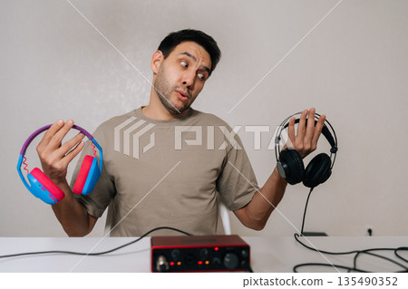 Portrait of confused excited man weighing colorful expressive headphones against black professional studio pair tied to audio interface, pondering audio quality, style, creative vs technical choice. Portrait of confused excited man weighing colorful expressive headphones against black professional studio pair tied to audio interface, pondering audio quality, style, creative vs technical choice. 135490352