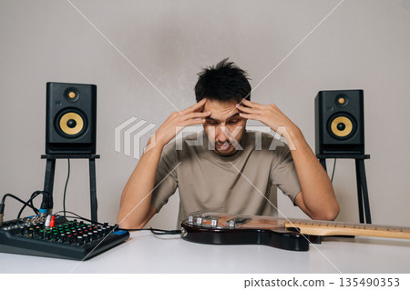 Portrait of frustrated man expressing confusion and uncertainty while sitting with audio equipment and bass guitar, struggling with music production and setup, at table on isolated white background. Portrait of frustrated man expressing confusion and uncertainty while sitting with audio equipment and bass guitar, struggling with music production and setup, at table on isolated white background. 135490353