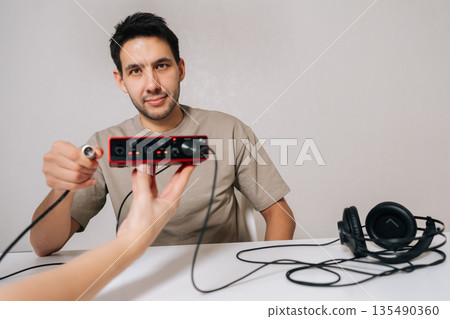 Man in beige t-shirt connecting xlr cable to compact red usb audio interface, preparing to use sound card equipment for music production, home studio setup or podcast recording. Man in beige t-shirt connecting xlr cable to compact red usb audio interface, preparing to use sound card equipment for music production, home studio setup or podcast recording. 135490360