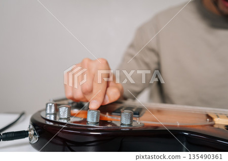 Close-up hands of musician adjusting sound, tone, volume on electric bass guitar with hand, preparing for performance or practice session in music studio or home setting, focusing on audio quality. Close-up hands of musician adjusting sound, tone, volume on electric bass guitar with hand, preparing for performance or practice session in music studio or home setting, focusing on audio quality. 135490361