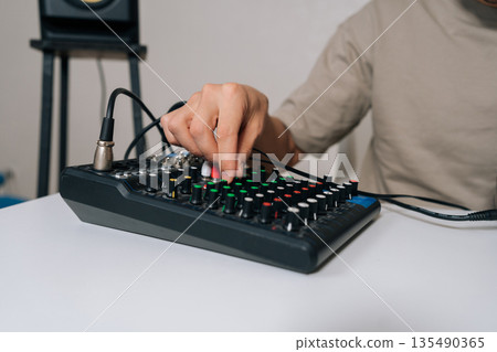 Close-up of person operating small audio mixing console, tuning input and output levels, balancing sound elements, mastering audio composition and delivering polished audio for listeners. 135490365