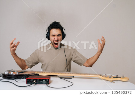 Portrait of emotional musician in headphones expressing intense anger and passion playing electric bass guitar in home studio setup with audio interface sitting at table on isolated white background. 135490372