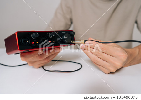 Person hands connecting audio jack cable into red usb audio interface, preparing for home music production, recording, and sound engineering in professional studio setup. Person hands connecting audio jack cable into red usb audio interface, preparing for home music production, recording, and sound engineering in professional studio setup. 135490373