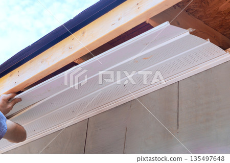 Worker installs white soffit panels on building while standing on ladder Worker installs white soffit panels on building while standing on ladder 135497648