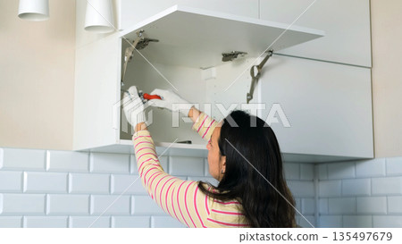 Young woman screws a screw and installs a shelf into a kitchen cabinet showing her skills during a home renovation project wearing gloves and staying focused on her task  135497679