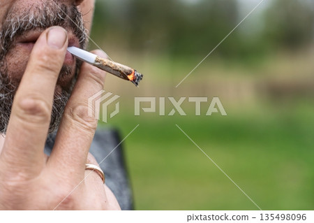 Bearded Man Smoking a Rolled Cigarette Outside with Blurred Green Background. Close-up View Bearded Man Smoking a Rolled Cigarette Outside with Blurred Green Background. Close-up View 135498096