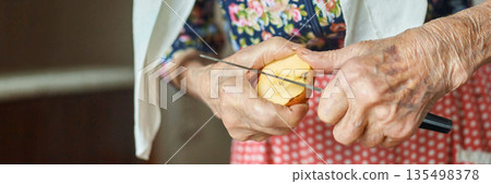 Elderly caucasian female peeling apple with knife in kitchen, floral blouse Elderly caucasian female peeling apple with knife in kitchen, floral blouse 135498378