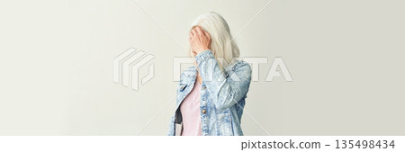 Elderly caucasian female in casual denim jacket covering face with hand against Elderly caucasian female in casual denim jacket covering face with hand against 135498434