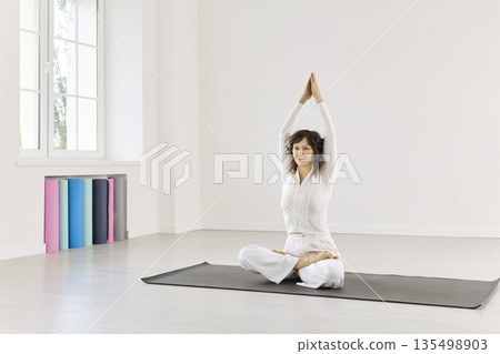 Serene indoor yoga practice: woman in lotus pose with raised arms on mat Serene indoor yoga practice: woman in lotus pose with raised arms on mat 135498903