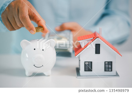 Hand holds coins over a white piggy bank beside a house model while cash is counted in the background, symbolizing mortgage down payment savings, real estate budgeting and smart finance. Hand holds coins over a white piggy bank beside a house model while cash is counted in the background, symbolizing mortgage down payment savings, real estate budgeting and smart finance. 135498979