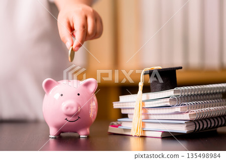 Hand puts a coin into pink piggy bank beside graduation cap and stacked notebooks on desk, symbolizing education savings, tuition budgeting, scholarship plans and smart student finance. 135498984