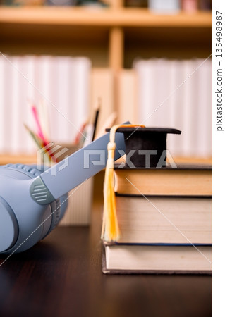 Gray headphones beside stacked textbooks with graduation cap and tassel on desk, blurred library background. Concept for online learning, audiobooks, study focus, education success. 135498987