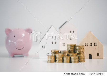 Wooden house model, coin stacks and pink piggy bank on white background, representing home savings, mortgage down payment, real estate budget planning and financial security goals. 135498990
