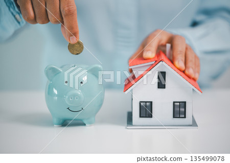 Hand drops a coin into a blue piggy bank beside a house model, symbolizing home savings, mortgage down payment, real estate budgeting and financial planning for buyers and future security. Hand drops a coin into a blue piggy bank beside a house model, symbolizing home savings, mortgage down payment, real estate budgeting and financial planning for buyers and future security. 135499078