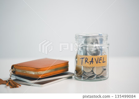 Glass jar labeled TRAVEL filled with coins and cash beside a leather wallet and notebooks on white background, symbolizing vacation savings, budget planning, travel fund goals and expenses. 135499084