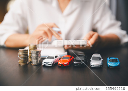 Stacks of coins beside a lineup of toy cars on a desk as a person calculates and writes notes, comparing car prices, auto loans, insurance costs and budgeting for a new vehicle purchase. 135499218