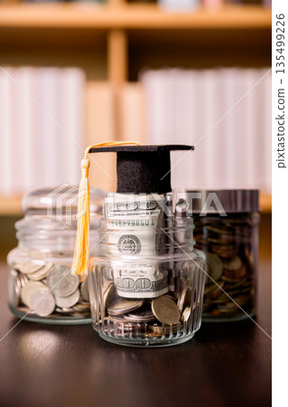 Graduation cap on a jar of coins and rolled dollar bills, with extra savings jars on desk and blurred bookshelf. Concept of tuition funding, education savings, budgeting and financial goals. Graduation cap on a jar of coins and rolled dollar bills, with extra savings jars on desk and blurred bookshelf. Concept of tuition funding, education savings, budgeting and financial goals. 135499226