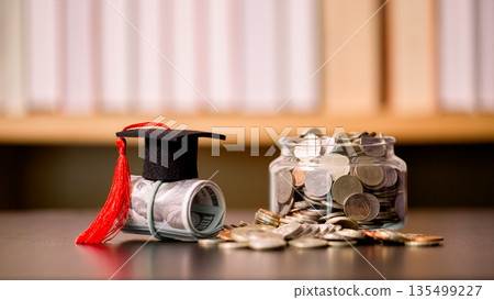 Graduation cap on a rolled banknote beside a glass jar of coins on a desk, symbolizing tuition savings, student budget planning, scholarships and financial goals for education funding and success. 135499227