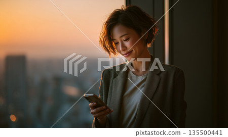 一位日本女性在城市建築內使用智慧型手機，背景是日落或夜景。這幅畫面展現了她的生活方式和溝通方式。 135500441