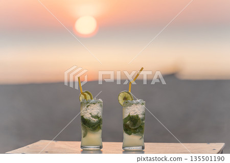 Two mojito cocktails on a table on the beach, against the background of the setting sun at sunset, sand and sea. The concept of relaxing and enjoying a vacation while traveling to exotic countries 135501190