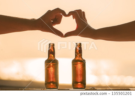 A couple in love drinks beer on the beach on Valentines Day. They make hearts out of their hands, depicting a beach, a honeymoon and a conceptual trip. They relax love each other and have fun on beach 135501201