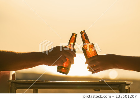 Two cold, steamed beer bottles in their hands clink glasses in close-up on the beach, against the background of the setting sun at sunset, sand and sea. Vacation and travel thirst quenching. High 135501207