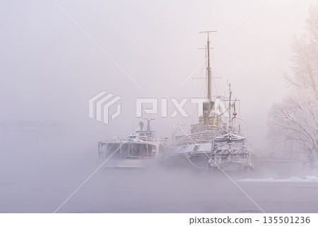 Old riverboat silhouettes are moored to a pier on the Yenisei River in heavy fog and frosty haze, creating an atmospheric nostalgic mood. Atmospheric river landscape in winter. High quality photo 135501236