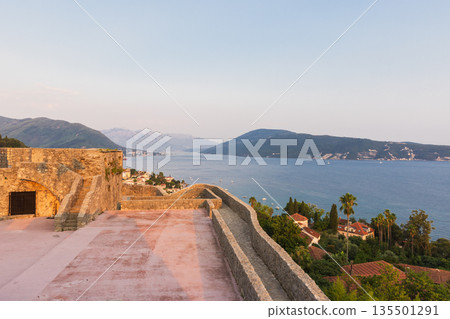 A historic stone terrace overlooks a tranquil bay with distant hills and a coastal town A historic stone terrace overlooks a tranquil bay with distant hills and a coastal town 135501291