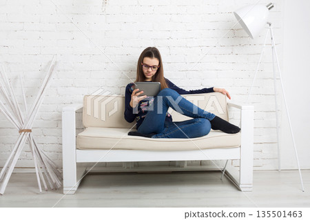 Teenage girl in eye glasses look into tablet, sitting on sofa. Children computer games, social networks and media addiction concept. Communication technologies, copy space 135501463