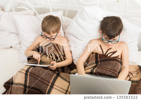 Boys in parents' bed at morning with laptop and tablet. Brothers play computer games. Siblings and gadgets. Children in glasses. Boys in parents' bed at morning with laptop and tablet. Brothers play computer games. Siblings and gadgets. Children in glasses. 135501522
