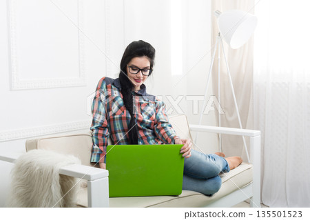 Freelancer woman works at home. Home office, freelance. Brunette girl with laptop in modern interior at white couch. High key. Freelancer woman works at home. Home office, freelance. Brunette girl with laptop in modern interior at white couch. High key. 135501523