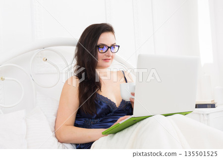 Morning coffee. Middle aged woman with laptop in bed. Beautiful woman in blue nightwear surfing internet at her computer. High key. 135501525