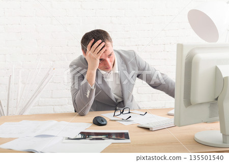 Working problems, issues of business. Young businessman in white modern office interior, thoughtful, sitting at computer table. Working problems, issues of business. Young businessman in white modern office interior, thoughtful, sitting at computer table. 135501540