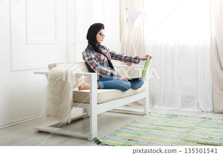 Girl with laptop works at home. Home office, freelance. Brunette in modern interior at white couch. High key. Girl with laptop works at home. Home office, freelance. Brunette in modern interior at white couch. High key. 135501541