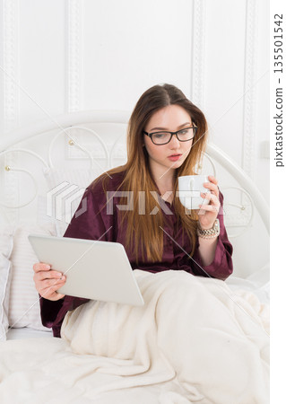 Sunday morning in bed. Young woman in surfing internet at her tablet pc and drinking coffee, copy space 135501542