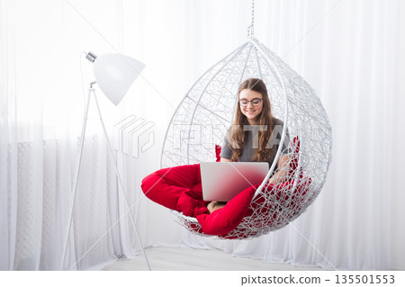 Beautiful woman in eyeglasses in fashionable armchair with laptop. Girl with pc in modern interior at red cushions. Stylish portrait of freelancer, work at home concept Beautiful woman in eyeglasses in fashionable armchair with laptop. Girl with pc in modern interior at red cushions. Stylish portrait of freelancer, work at home concept 135501553