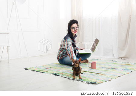 Young girl in computer glasses on carpet with laptop and dog chihuahua. Hipster freelancer woman works at home. Home office, freelance. Young girl in computer glasses on carpet with laptop and dog chihuahua. Hipster freelancer woman works at home. Home office, freelance. 135501558