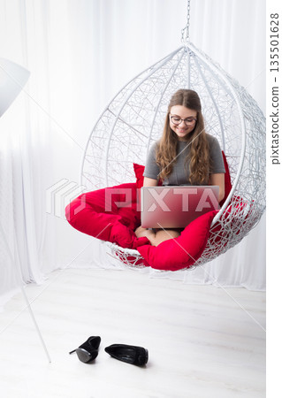 Beautiful woman in eyeglasses in fashionable armchair with laptop. Girl with pc in modern interior at red cushions. Stylish portrait of freelancer, work at home concept 135501628