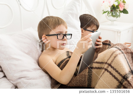 Boys in parents' bed at morning with laptop and tablet. Brothers play computer games. Siblings and gadgets. Children in glasses. 135501656