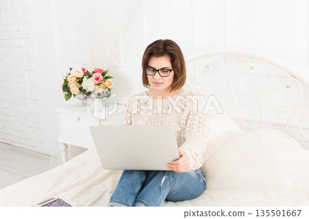 Young woman freelancer works at home in bed with laptop. Engrossed Girl in eyeglasses. High key. Freelance. 135501667