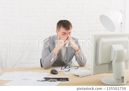 Overworking, toried at work. Young businessman in white modern office interior, sitting at his desk Overworking, toried at work. Young businessman in white modern office interior, sitting at his desk 135501730