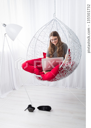 Beautiful woman in eyeglasses in fashionable armchair with laptop. Girl with pc in modern interior at red cushions. Stylish high key portrait of freelancer, work at home concept. Beautiful woman in eyeglasses in fashionable armchair with laptop. Girl with pc in modern interior at red cushions. Stylish high key portrait of freelancer, work at home concept. 135501732