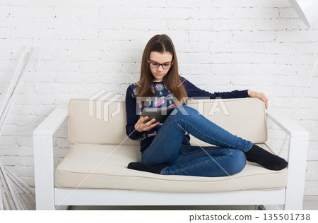 Teenage girl in eye glasses look into tablet, sitting on sofa. Children computer games, social networks and media addiction concept. Communication technologies, copy space 135501738