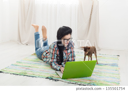 Young girl in computer glasses lying on carpet with laptop and dog chihuahua. Hipster freelancer woman works at home. Home office, freelance. Young girl in computer glasses lying on carpet with laptop and dog chihuahua. Hipster freelancer woman works at home. Home office, freelance. 135501754