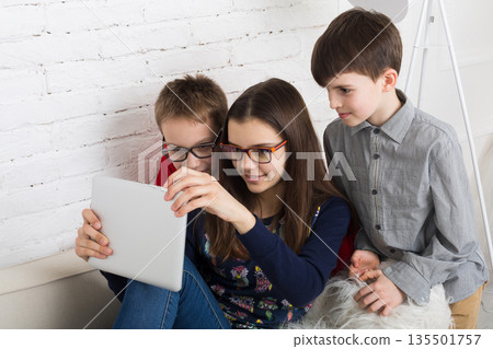 Group of kids in eye glasses look into tablet. Children computer games, social networks and media addiction concept. Girl and boys with gadget. Communication technologies. 135501757