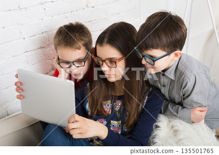 Group of kids in eye glasses look into tablet. Children computer games, social networks and media addiction concept. Girl and boys with tablet. Communication technologies. 135501765